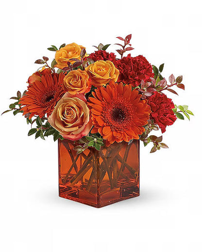 Colorful flower arrangement with orange, red, and yellow flowers in a transparent orange vase on a white background.