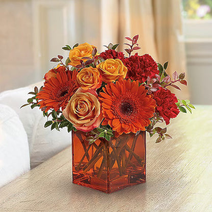 Bouquet of orange, red, and yellow flowers in a clear vase on a wooden surface.