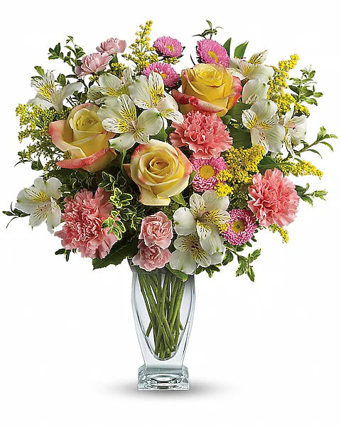 A bouquet of mixed flowers in a clear glass vase, featuring yellow roses, pink carnations, white alstroemeria, pink asters, and sprigs of yellow solidago