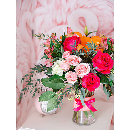 Bouquet of mixed flowers in a clear glass vase tied with a pink ribbon against a pink flamingo-themed background.